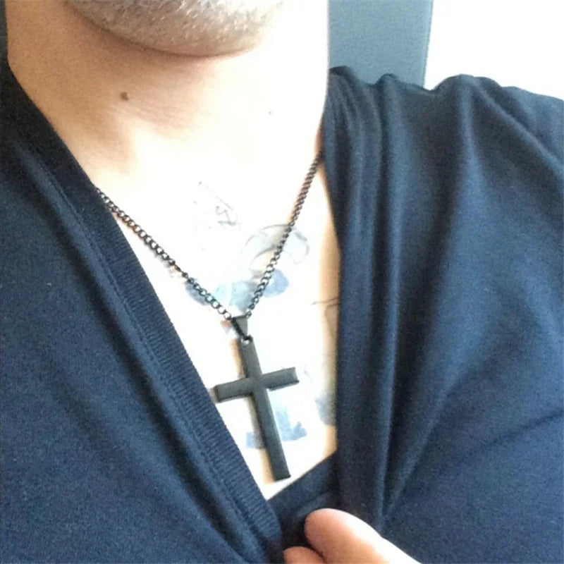 Man wearing a black cross pendant necklace over a tattooed chest and dark shirt